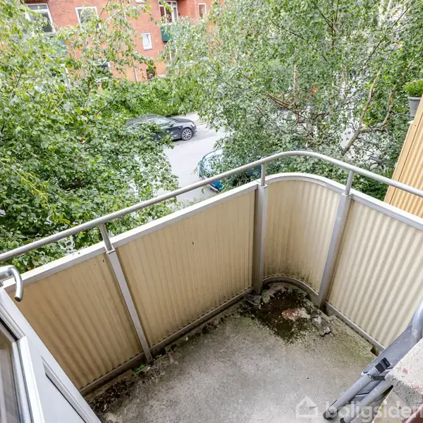 Balkon med metalgelænder virker rolig, omgivet af frodige grønne træer. En åben dør fører ud til balkonen, som har udsigt over en gade med parkeret bil og røde murstensbygninger.
