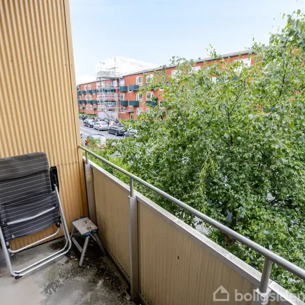 En altan med sammenklappelige stole og et bord står stille. I baggrunden ses grønne træer og røde murstensbygninger langs en vej.