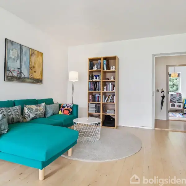 Turkis sofa med puder står i en lys stue ved siden af et hvidt rundt bord på et tæppe. Bogreol fyldt med bøger ses ved siden af en gulvlampe.