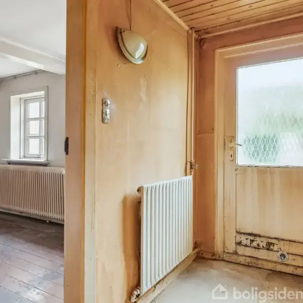 En trægulvet entré med et slidt dørparti ved siden af en radiator leder ind til et lyst rum med vinduer og radiatorer. Veggene er træpaneler med en væglampe.
