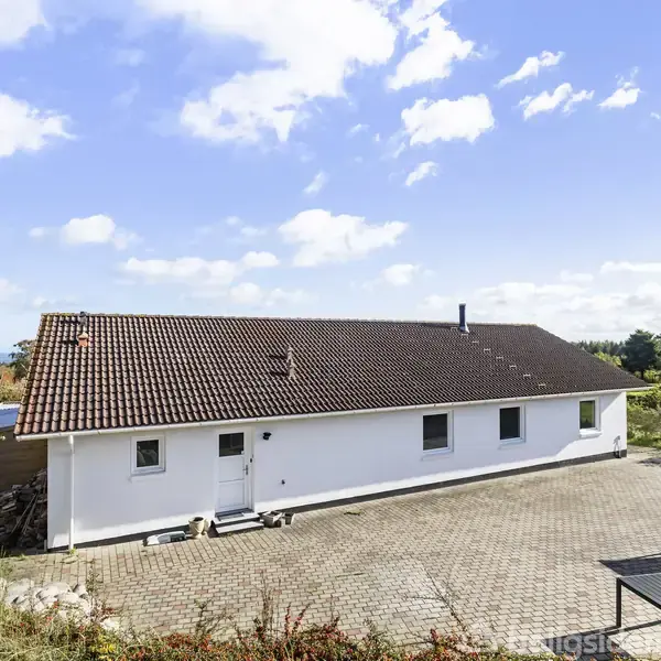 En hvidmalet bygning med rødt tag står stille ved en stenbelagt gårdsplads i et roligt forstadsområde, omgivet af grønne træer og med udsigt til en klar, blå himmel.