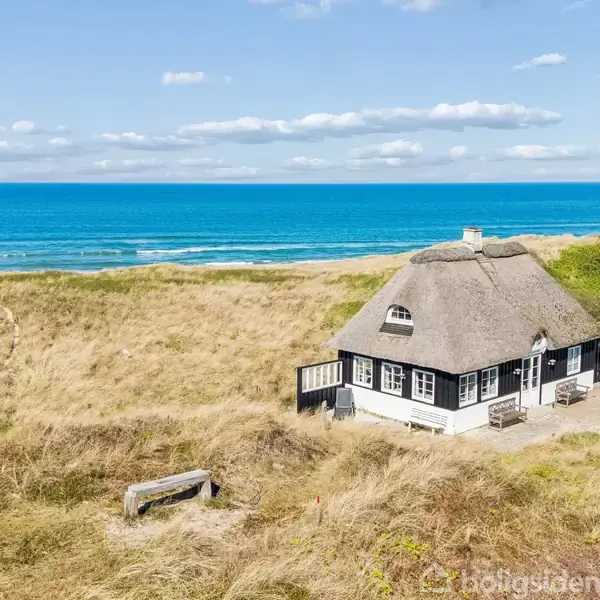 Stråtækt hus står stille i klitlandskab ved kysten. Det ligger tæt på havet med blå bølger og blå himmel i baggrunden, omgivet af gyldent græs og buske.