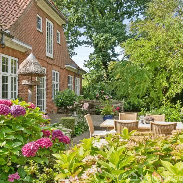 En murstensbygning med store vinduer står ved siden af en terrasse, hvor der er havemøbler. Omgivet af blomstrende hortensiaer og frodige grønne træer under en klar himmel.