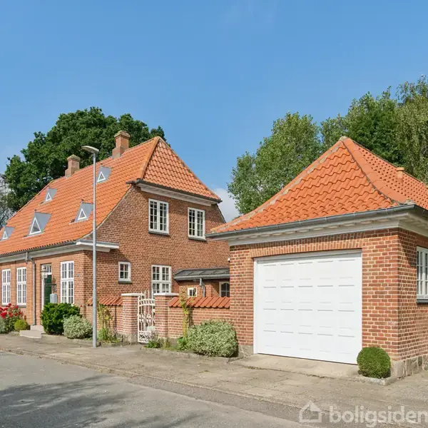 Rødt murstenshus med røde tegltag, omgivet af træer og have. Til højre en tilhørende garage. Husets facade har flere vinduer, og det ligger ved en stille vej.