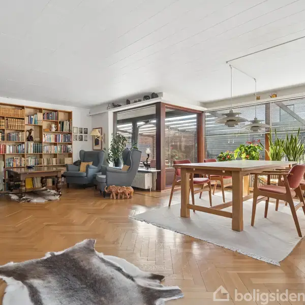 Stue med bogreol fyldt med bøger, to lænestole ved et lille sofabord. Spisebord med seks stole ligger på tæppe nær vinduer, der giver udsigt til haven. Hvidt loft.