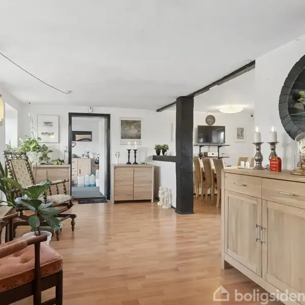 Kommode med dekorationer står langs en væg i et åbent opholdsrum. Rundt spejl hænger over kommoden. Stueplanter og stole tilfører hygge, mens spisestuestole ses i baggrunden.