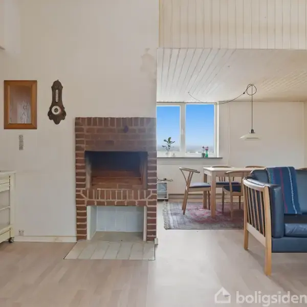 En stue med en murstenspejs står stille ved væggen. Til venstre ses et serveringsbord, mens til højre er et område med sofa og spisebord, oplyst af vindueslys.