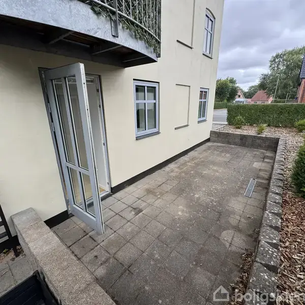 En åben dør i en bygning leder ud til en flisebelagt terrasse. Terrassen er omgivet af lave mure og en hæk, med et hus og træer i baggrunden.