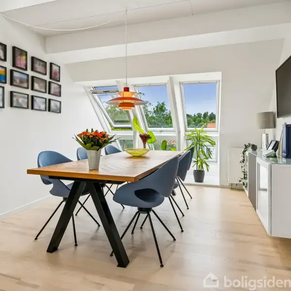 Spisebord med seks blå stole står i et lyst rum. En orange lampe hænger over bordet, blomsterdekorationer pynter. Væggene har billedrammer, og store vinduer viser grøn udsigt.