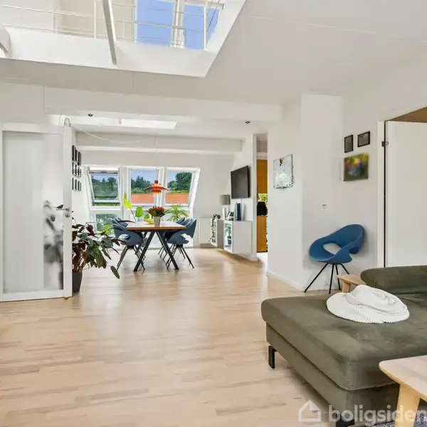 Stue med grøn sofa og sofabord, åbner op til spisestue med spisebord og stole, lyst trægulv, masser af naturligt lys fra vinduer og ovenlysvinduer, dekoreret med malerier.