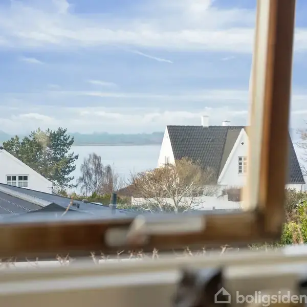 Et åbent vindue viser udsigten til et hvidt hus og havet i baggrunden. Tager sted på en solrig dag med blå himmel og skyer.
