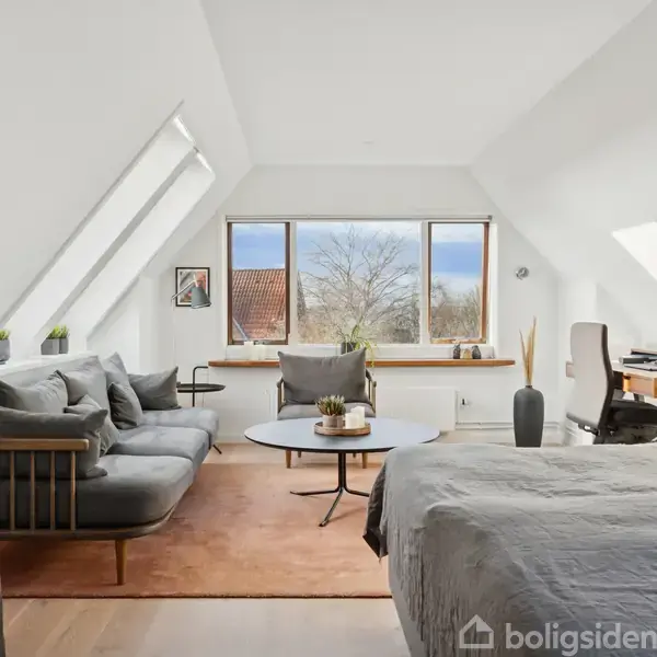 En lys stue med en grå sofa og sofabord til venstre, en seng i forgrunden, skråvinduer, og et skrivebord bagved. Store vinduer viser et rødtaget hus og træer udenfor.