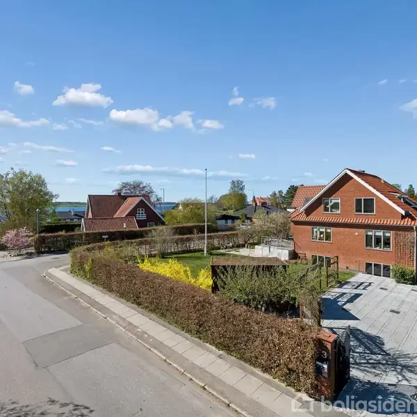 Rødt murstenshus står på en solrig vej omgivet af andre huse og grønne haver. Huset har en flisebelagt indkørsel, og træer er synlige i baggrunden.