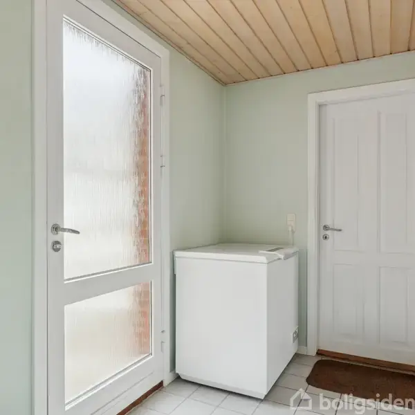 En hvid kummefryser står i et hjørne mellem en matglasset dør og en lukket dør i et rum med lys trægulv og træloft. En lille firkantet spejl hænger på væggen.