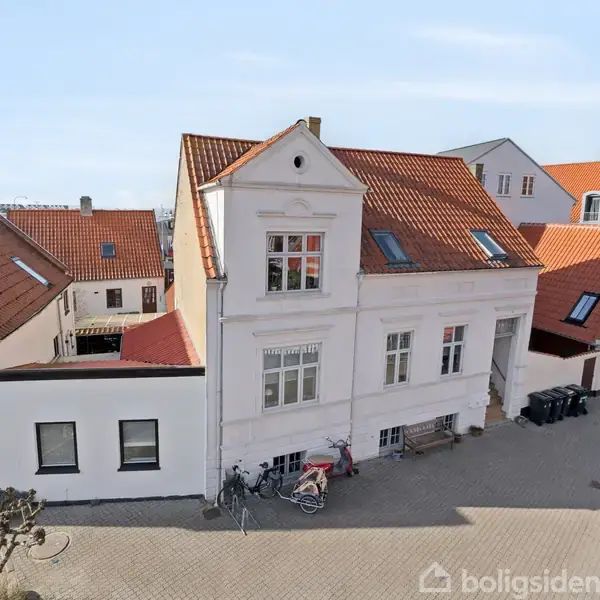 Hvidt, ældre hus med rødt tegltag står stille i en brolagt gårdsplads. Cykler er parkeret foran, og andre lignende huse omkranser området.