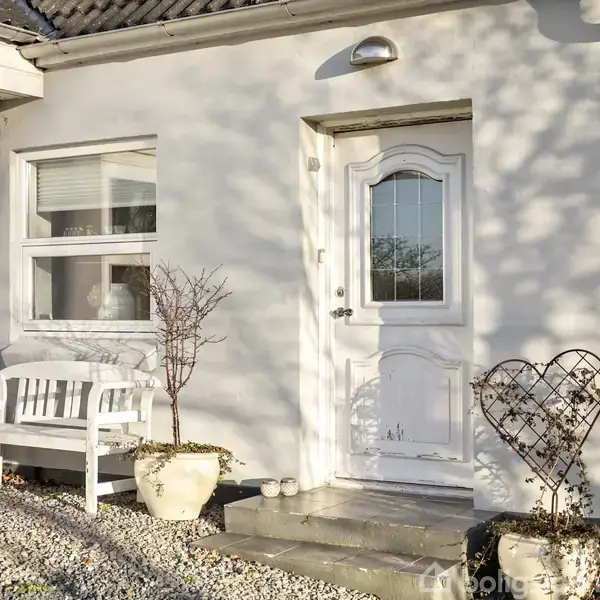 En hvid hoveddør med glaspanel er omgivet af to potteplanter. En hvid bænk står ved siden af, mens hjertedekorationen af metal er placeret nær indgangen. Grus dækkede jorden.