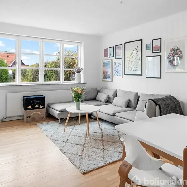 En lys, moderne stue med en grå hjørnesofa, sofabord og vægkunst. Store vinduer giver udsigt til en have. Et spisebord og stole står ved siden af.