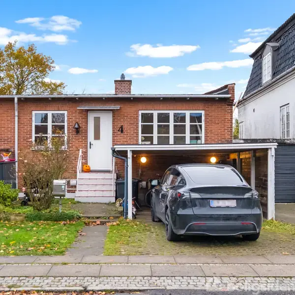 Rød murstenshus med hvide vinduer, beliggende i en forstadsgade. En bil holder parkeret under en carport, omgivet af efterårsblade på græsset.