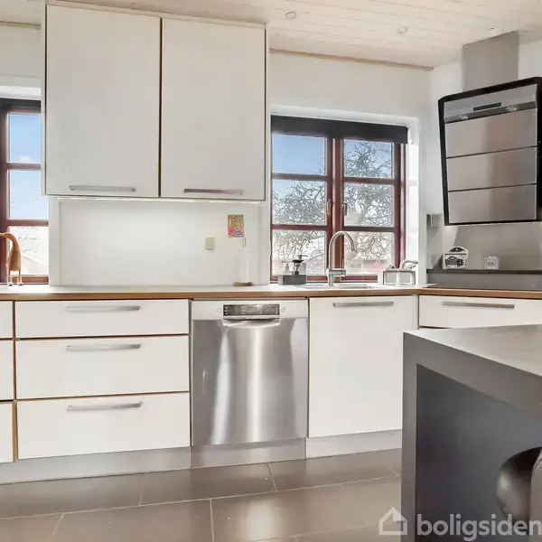 Hvidt, moderne køkken med træbordplader og to vinduer med udsigt til træer. Der er opvaskemaskine, kaffemaskine og køkkenredskaber på bordet i et lyst rum.