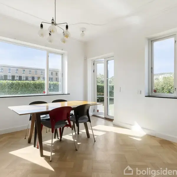 Spisebord med stole står i et lyst rum med trægulv, omgivet af store vinduer med udsigt til en have. Et moderne loftlys hænger over bordet.