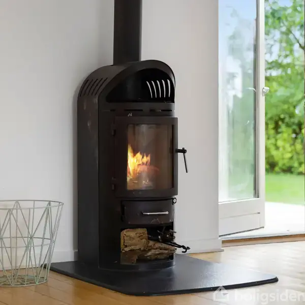 En brændeovn, der brænder træ, står i en lys stue ved siden af en åben dør med udsigt til en grøn have. Der er også en metaltrådkurv ved siden af ovnen.