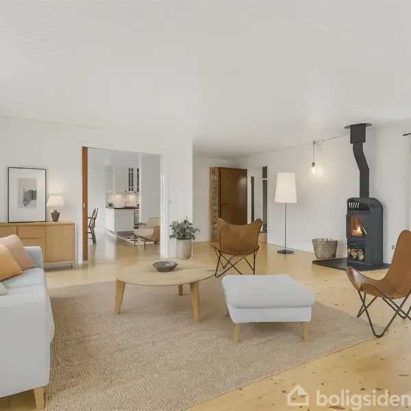 En stue med en lys sofa og to læderstole rundt om et rundt sofabord. En brændeovn er tændt, og rummet har lyse trælofter og gulve, der skaber en hyggelig atmosfære.