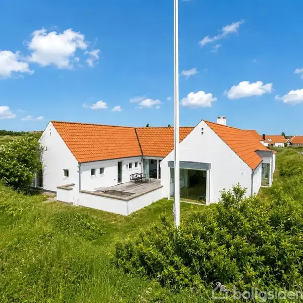 Et hvidt hus med rødt tegltag står på en grøn bakke nær en vej. I baggrunden er der flere huse og en blå himmel med skyer.
