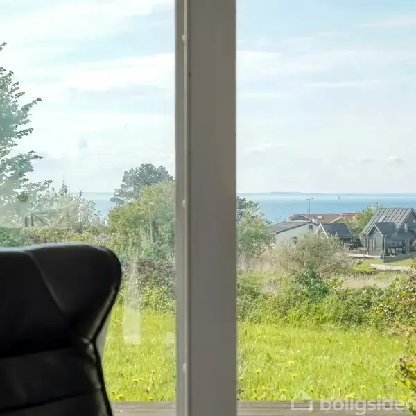 En lænestol står ved et vindue, der viser udsigt over en grøn græsplæne, huse og havet i horisonten.