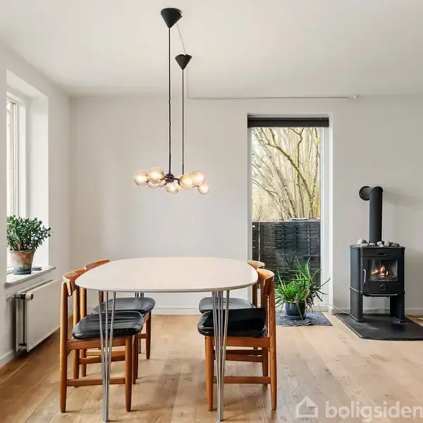 Et spisebord med fire stole står i et lyst rum med en moderne loftslampe over. Til højre er der en brændeovn ved siden af en dør, der fører udenfor.