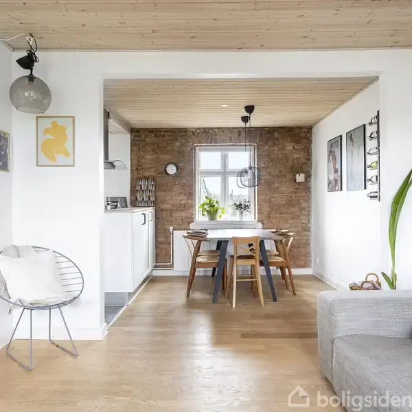 Rummet har en grå sofa og en hvid stol, stående mod en hvid væg. Et spisebord med stole ses i baggrunden foran en murstensvæg. Der er planter og kunst på væggene.