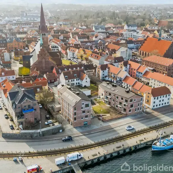 Kirke med højt spir står centralt omgivet af røde tage i en tæt bymidte nær en havn med fiskerbåde langs kajen. bymiljø.