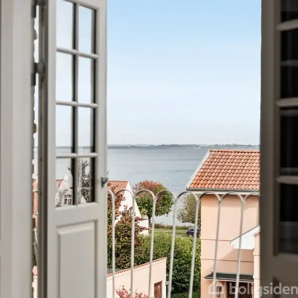 Dobbeltfranske døre står åbne mod en balkon med udsigt til havet. Røde tegltage og grønne træer ses i det omkringliggende landskab.