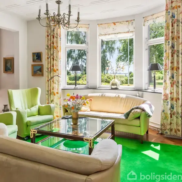 Stue med lysegrønne stole og en beige sofa omkring et glasbord med blomster i vase. Store vinduer med blomstrede gardiner giver udsigt til en frodig have. En lysekrone hænger fra loftet.