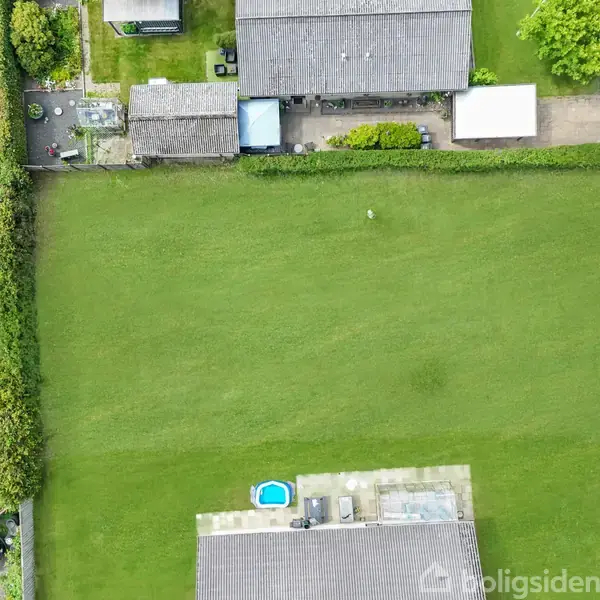 Græsplæne omkranset af hække, set fra oven, med en lille pool og terrasse i nederste del; flere huse og en vej i baggrunden.