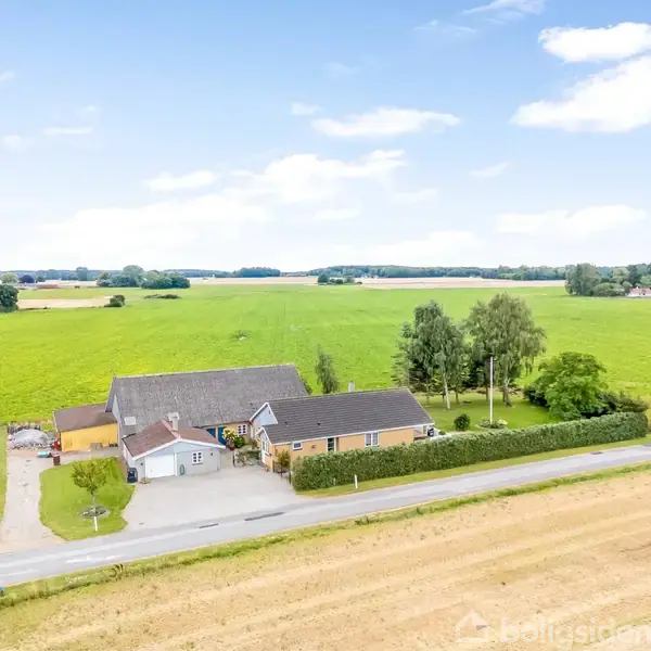 Et hus ligger stille på en landejendom omgivet af grønne marker under en blå himmel med få skyer. Flere træer er synlige tæt på huset, som er af gult murværk.