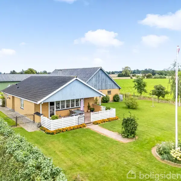 Et enkelt enfamiliehus med sort tag og gule ydermure står stille omkranset af grønne træer og en velplejet græsplæne i et åbent landligt område under en blå himmel.