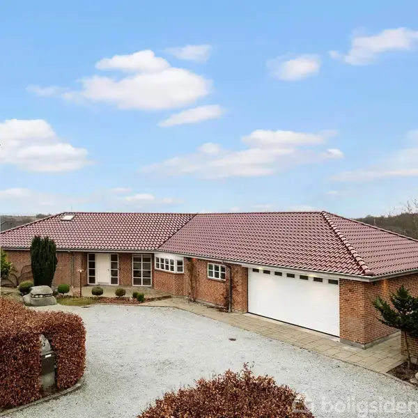 En murstensvilla med rødt tegltag står centralt på billedet. Indkørslen er grusbelagt, omkranset af hæk, og landskabet er åbent med klar himmel og spredte træer i baggrunden.