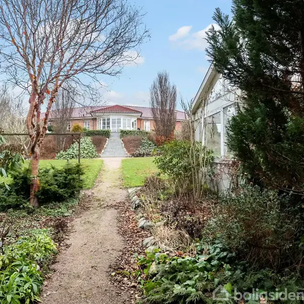 En bred sti fører op til et hus omgivet af træer og buske. Haven er frodig, med græs og forskellige planter, og himlen er blå med spredte skyer.