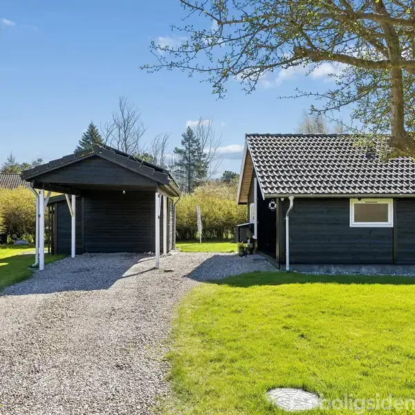 Sort træhus med grå fliser står ved siden af en carport af samme materiale. Grusvej leder til huset, omgivet af grønt græs og træer under en blå himmel.