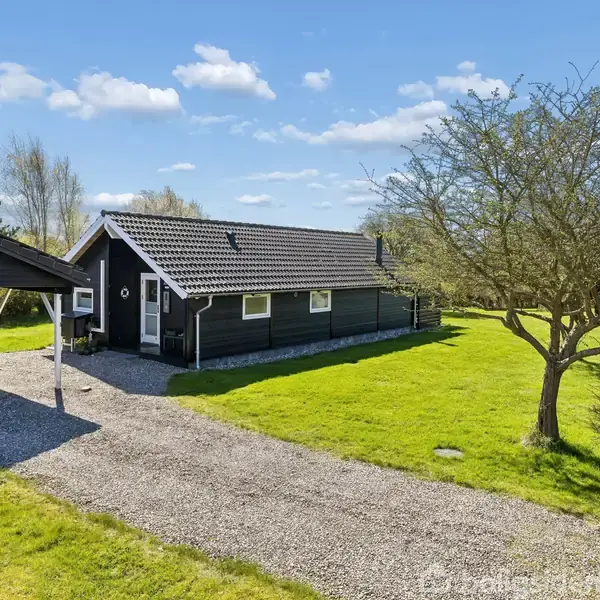 Et sort sommerhus står på en grøn græsplæne med en grussti foran. En carport er til venstre, og der er et træ ved siden af.