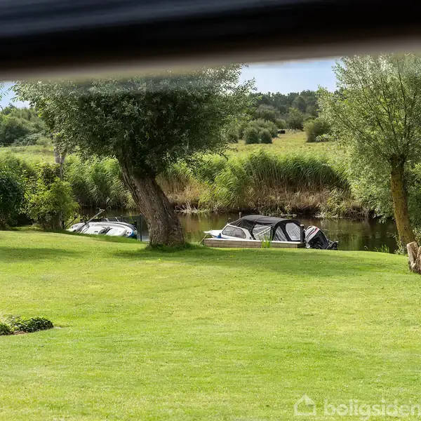 Både ligger stille ved bredden af en sø, omgivet af grønne græsplæner og træer, set gennem en vinduesramme.