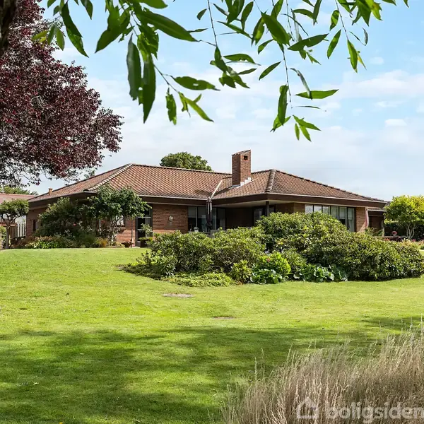 Et rødt murstenshus med en brun tegltag står omgivet af en velplejet have med grønne buske og træer. Der hænger grene i forgrunden under en blå himmel.
