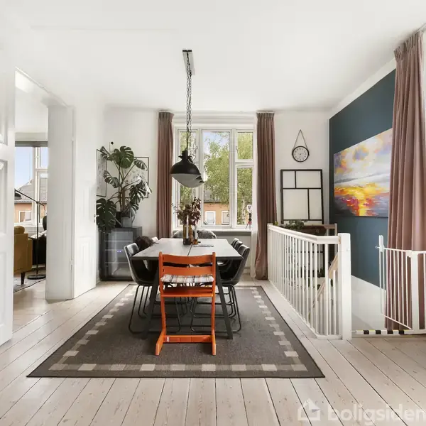 Spisebord med stole står på tæppe i spisestue. Rummet er lyst med planter, en stor lampe, maleri på væggen og udsigt til grønne træer udenfor vinduerne.