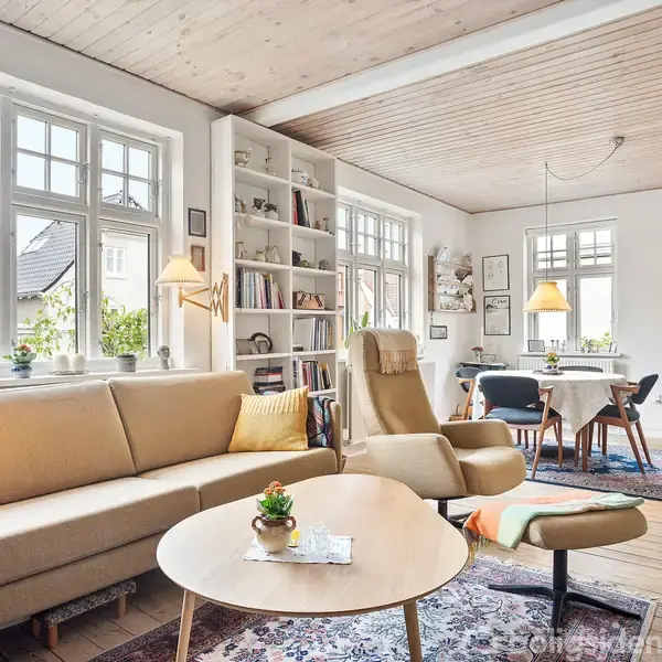 Stue med lyse møbler, herunder en sofa og lænestol foran et rundt sofabord. Der er reoler fyldt med bøger. Store vinduer slipper lys ind. Ved bordet ses stole og lamper.