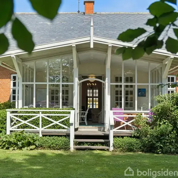 Hus med skråtag og glasveranda. Terrassen har hvide hegn og er omgivet af grønne planter. Indretningen indenfor kan svagt ses gennem døren. En have omgiver huset.