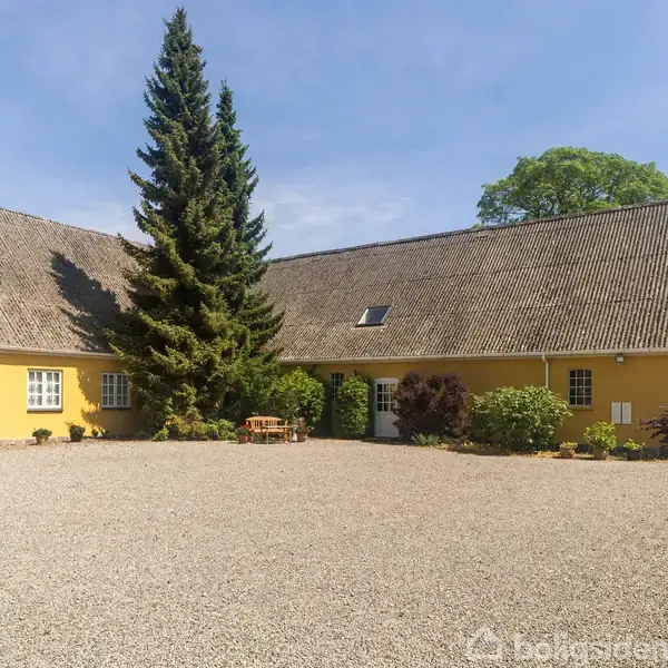Gult bondehus med stor gruset gårdsplads, omgivet af buske og et højt grantræ. Huset har flere hvide vinduer, og rundt om er der en lysblå himmel.