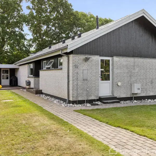 En lys murstensvilla med sort tag ses fra siden, omgivet af en velplejet græsplæne og en flisebelagt sti. Tæt bevoksning grænser op til grunden.