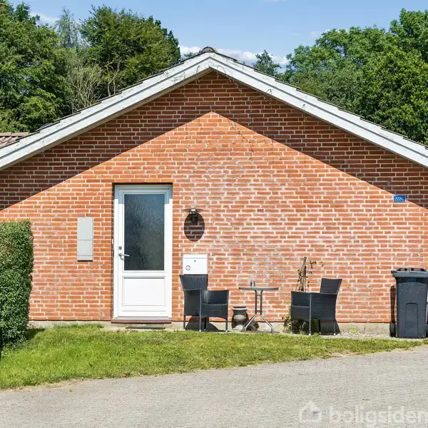 Rødt murstenshus med hvid dør, to sorte stole og bord ved indgangen. Flere træer i baggrunden. En affaldsbeholder står til højre. Himmel med spredte skyer ses ovenfor.