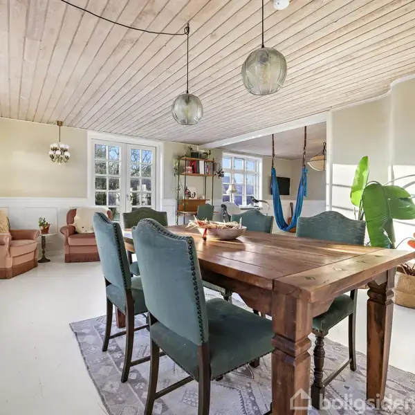 En spisestue med et solidt trækbord centralt placeret, omgivet af seks polstrede stole. Store vinduer og lyse vægge skaber en lys atmosfære, suppleret af grønne planter og moderne lamper.