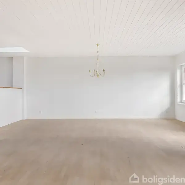 En tom stue med en lys trælaminatgulv, hvide vægge og loft. En simpel lysekrone hænger fra loftet. Store vinduer giver dagslys, der oplyser rummet.